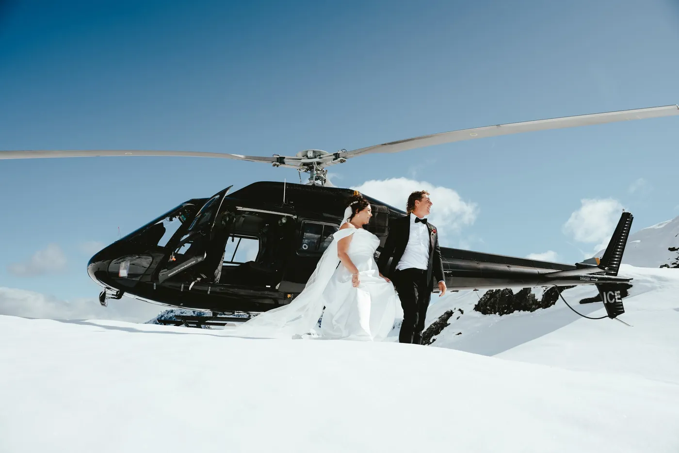 A bride and groom in wedding attire standing beside a black helicopter on a snowy mountain, with snow-covered peaks and a blue sky in the background.